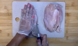 Person slicing raw chicken breasts on a cutting board with knife, wearing gloves for hygiene.